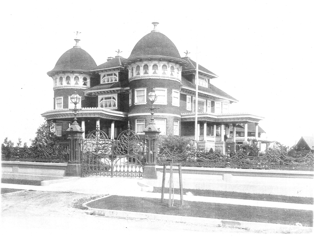 Historic black and white photo of the Canuck Place Vancouver hospice, a majestic Victorian mansion featuring multiple rounded towers and ornate architectural details. The house is surrounded by an elaborate wrought iron fence with intricate designs. This grand building, showcasing expansive porches and multiple stories, is set in a serene, well-manicured landscape.