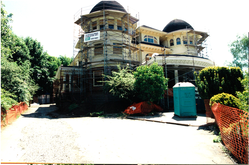 Canuck Place Vancouver hospice, a Victorian mansion undergoing renovations, wrapped in scaffolding and construction materials. The mansion features twin towers and intricate architectural details, currently obscured by the construction work. In the foreground, construction barriers and a portable toilet indicate an active work site. The surrounding area includes mature trees and bushes.