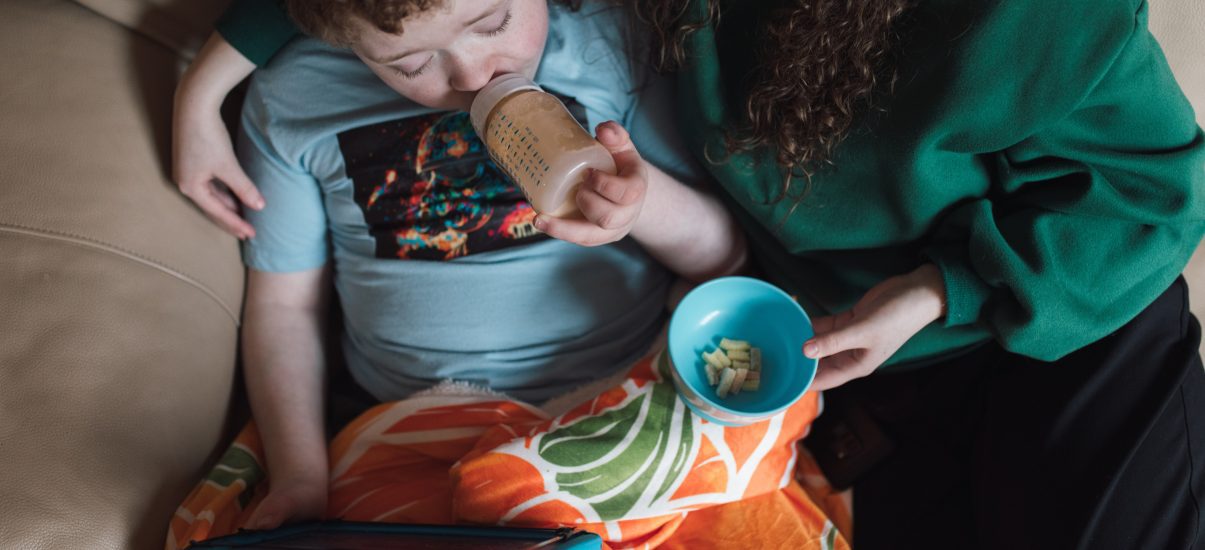 A mother sits on a couch with her young child, who is drinking from a bottle and watching something on an iPad. The mother is handing the child some food, creating a cozy and nurturing atmosphere as they share this moment together.