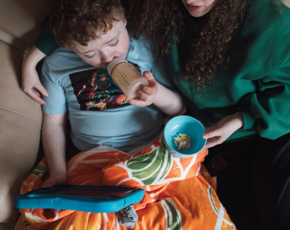 A mother sits on a couch with her young child, who is drinking from a bottle and watching something on an iPad. The mother is handing the child some food, creating a cozy and nurturing atmosphere as they share this moment together.