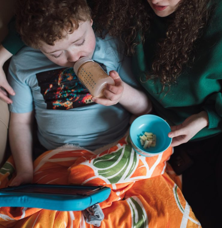 A mother sits on a couch with her young child, who is drinking from a bottle and watching something on an iPad. The mother is handing the child some food, creating a cozy and nurturing atmosphere as they share this moment together.